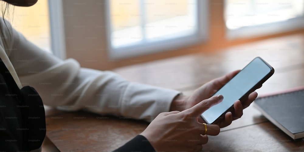 Cropped image of stylish woman's hands holding a white blank screen smartphone while sitting and relaxing at the wooden working desk over comfortable living room windows as background.