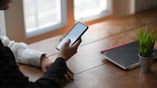 Cropped image of stylish woman's hands holding a white blank screen smartphone while sitting and relaxing at the wooden working desk over comfortable living room windows as background.