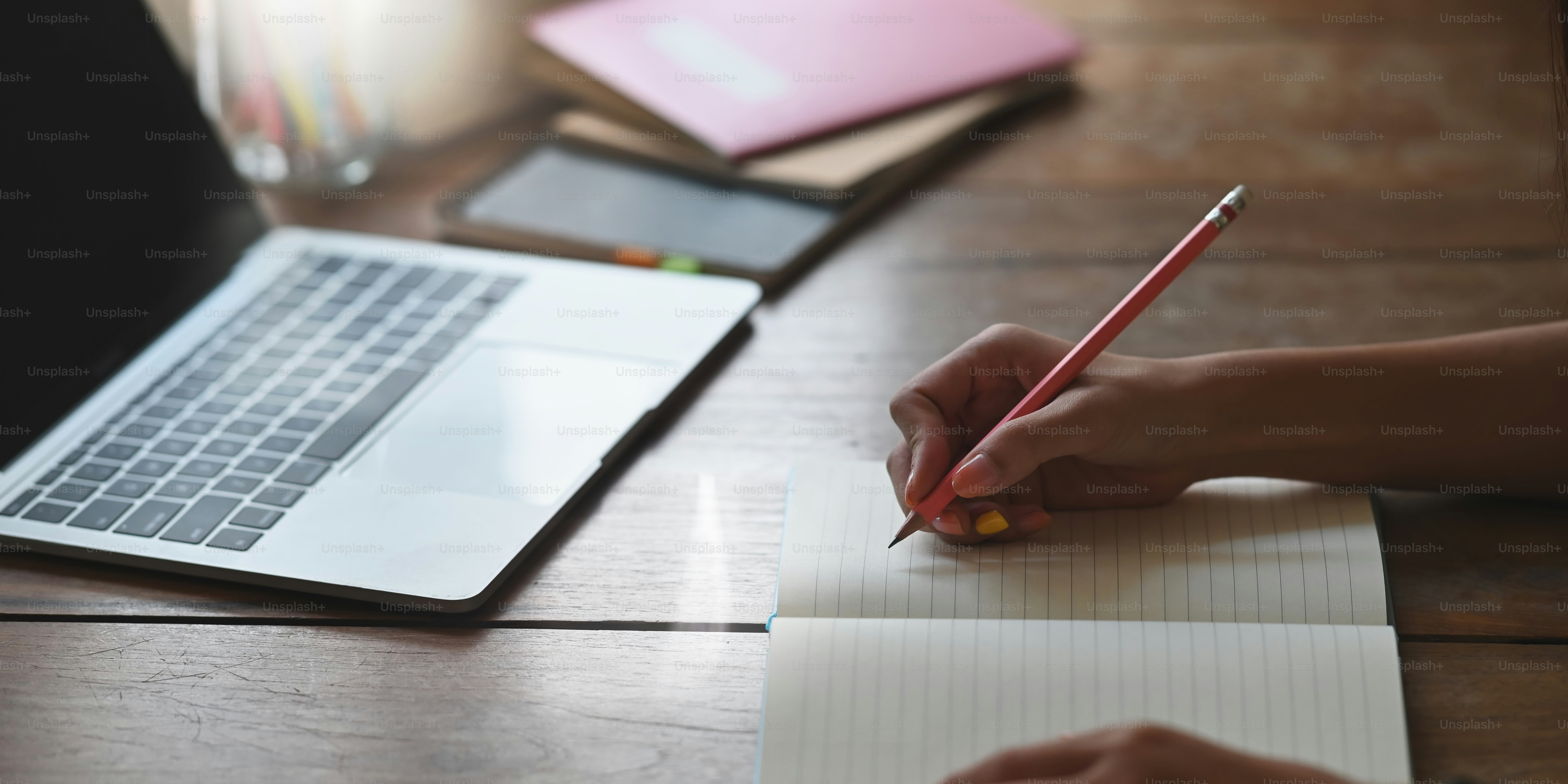 Imagen recortada de una hermosa mujer escribiendo en un cuaderno que se pone en el escritorio de trabajo y rodeada de computadora portátil y cuaderno sobre una cómoda sala de estar como fondo.