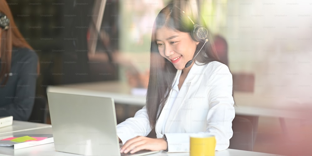 Beautiful woman looks good in white suit working as call center/operator/customer call service while sitting at the white working desk over her colleagues in modern  office as background.