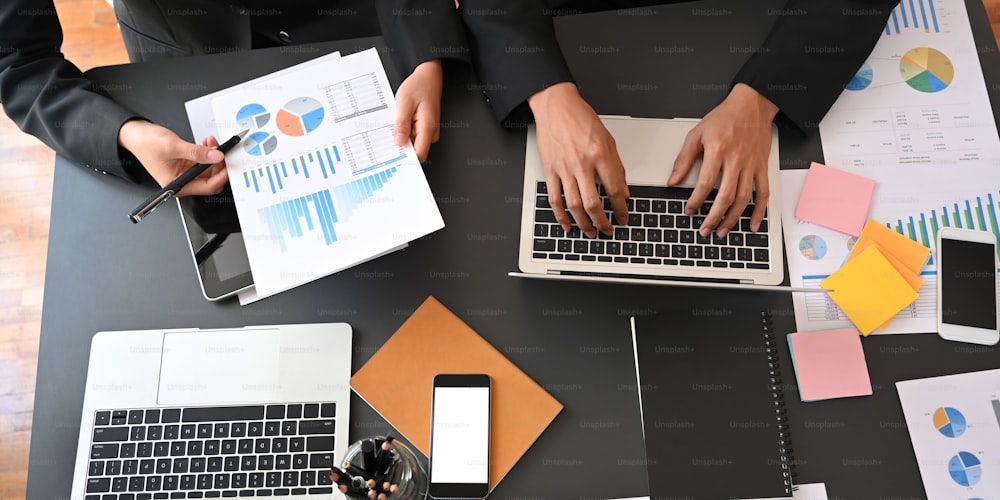 Top view of business executive men discussing about their business plan with computer laptop and graphic charts while sitting together at black meeting table over modern office as background.