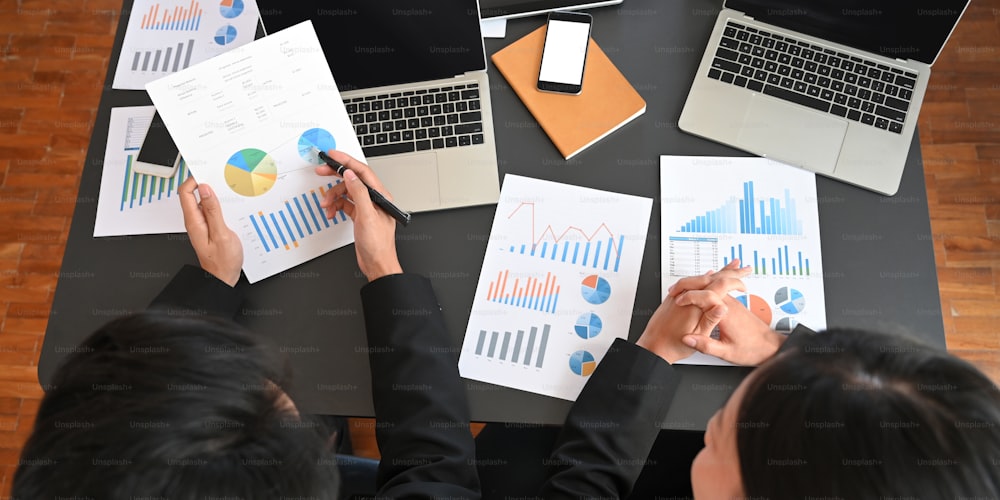 Top view of business executive people discussing about their business plan with computer laptop and graphic charts while sitting together at black meeting table over modern office as background.