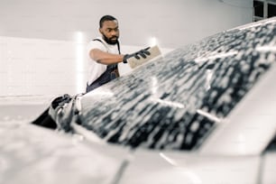 Young African man, car wash worker, wearing t-shirt and overalls cleaning the windshield of car with the help of special foam and sponge. Washing the car by hand, sponge and foam bubbles.