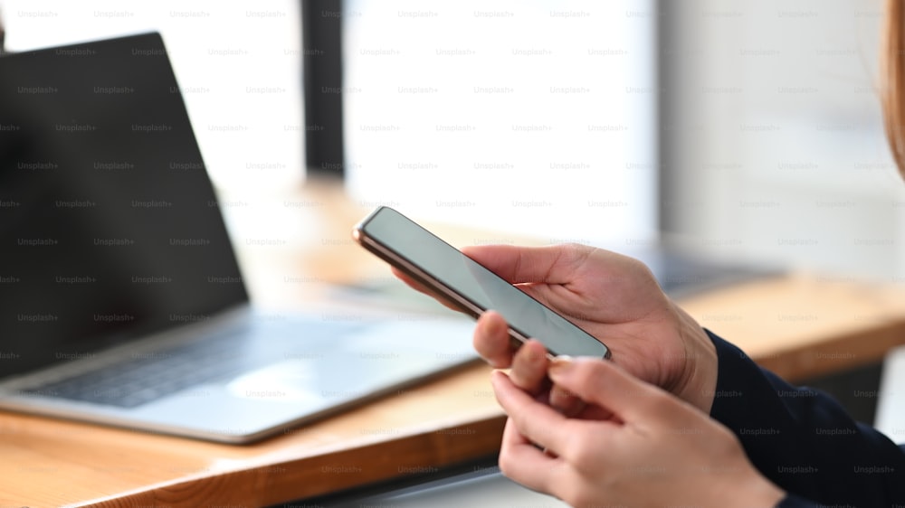 Image recadrée de la main d’une femme d’affaires tenant un smartphone sur un ordinateur portable mettant sur un bureau de travail en bois comme arrière-plan. Concept de technologie et de style de vie.