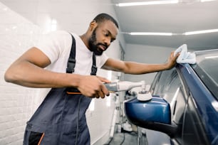 Car polish wax and detailing. Young male African worker, wearing white t-shirt and gray overalls, holding a polisher and polishing car at professional car detailing service.