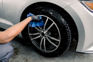 Car rims cleaning, car detailing wash concept. Cropped close up photo of male hand in black rubber glove with blue microfiber cloth washing car alloy wheel at car wash service.