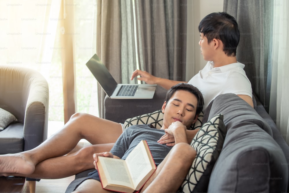 Asian homosexual gay man couple using laptop working at home with boyfriend relaxing on sofa in living room.