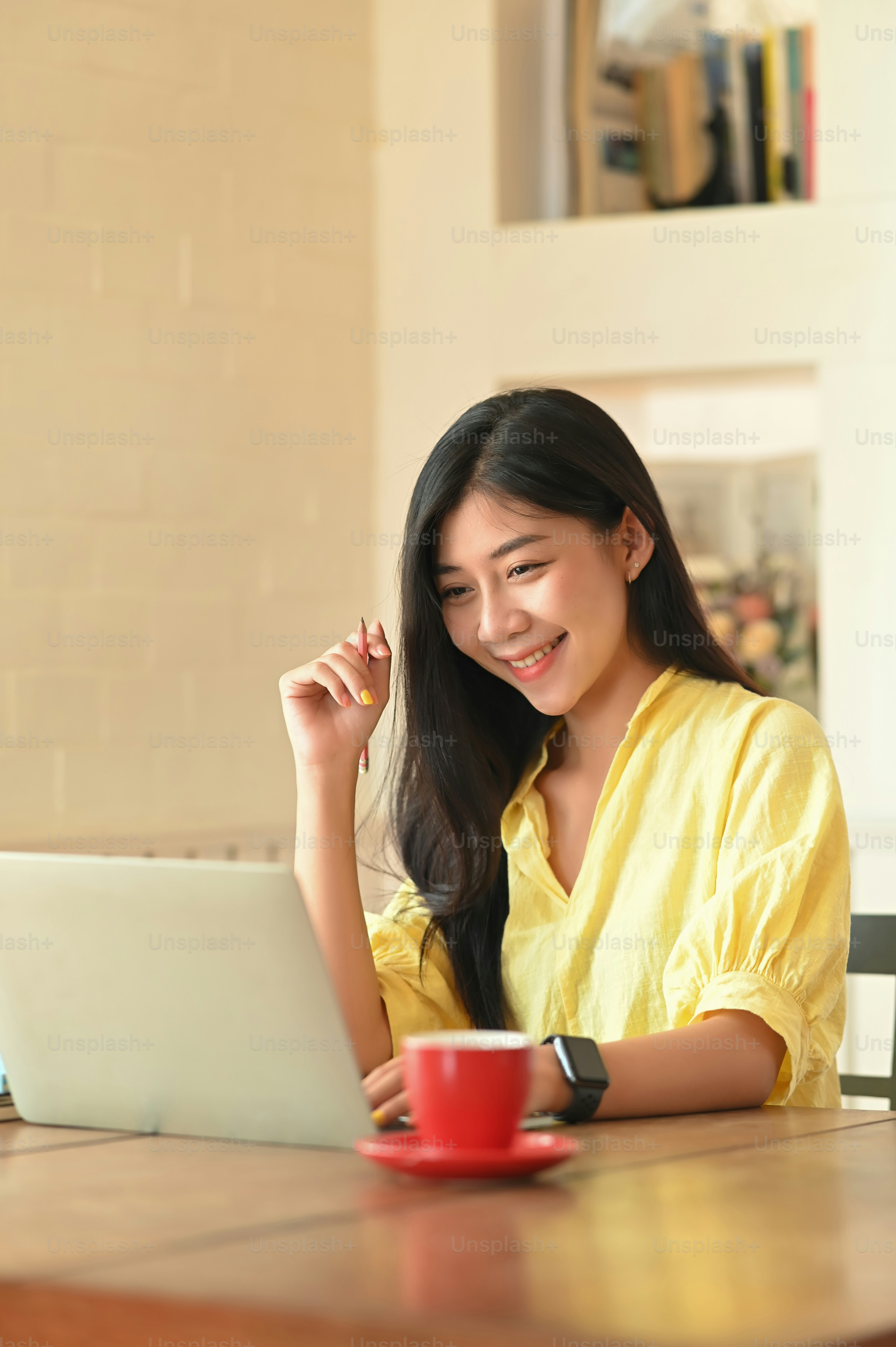 Foto de una mujer hermosa que se ve bien con una camisa de algodón amarilla sonriendo y mirando fijamente la computadora portátil mientras está sentada en el escritorio de trabajo de madera sobre una cómoda sala de estar como fondo.