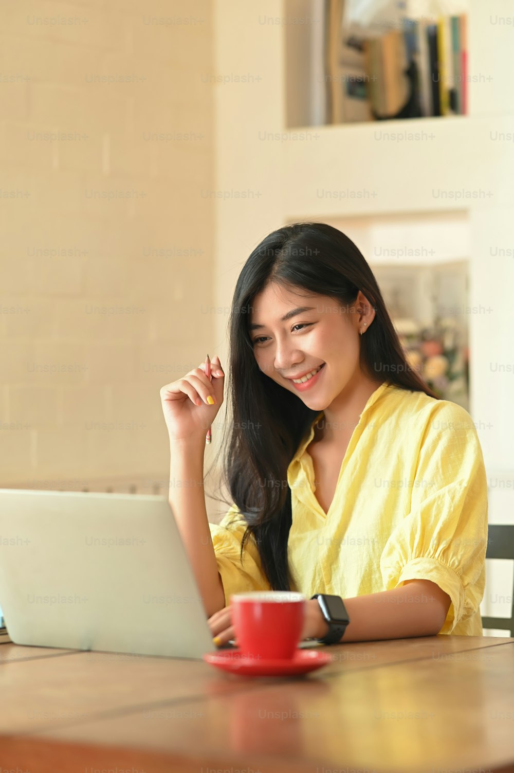 Photo of beautiful woman that look good in yellow cotton shirt smiling and staring at the computer laptop while sitting at the wooden working desk over comfortable living room as background.