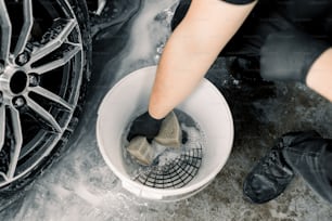 Concept de lavage de voiture et d’esthétique. Vue de dessus des outils de lavage dans le service de lavage de voiture, seau blanc avec solution de nettoyage au savon, grille spéciale. Main d’un ouvrier masculin tenant une éponge pour nettoyer les jantes de voiture.