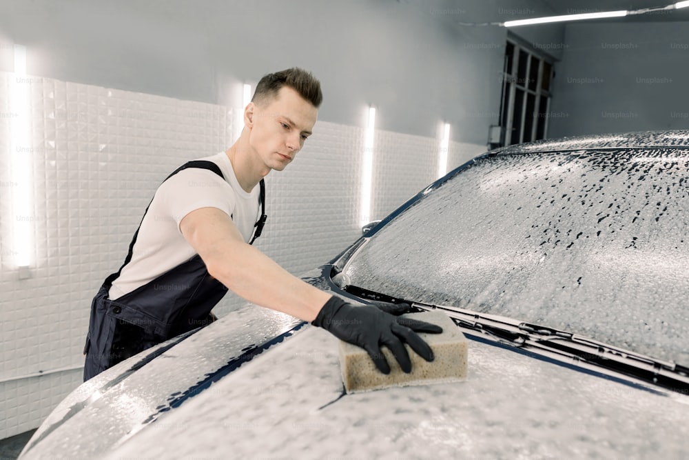 Giovane operaio maschio di servizio dell'automobile che pulisce il cofano dell'automobile con l'aiuto di shampoo e spugna gialla nell'azienda di lavaggio auto.