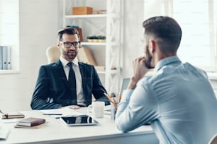 Zwei junge Männer in formeller Kleidung kommunizieren miteinander, während sie im Büro sitzen