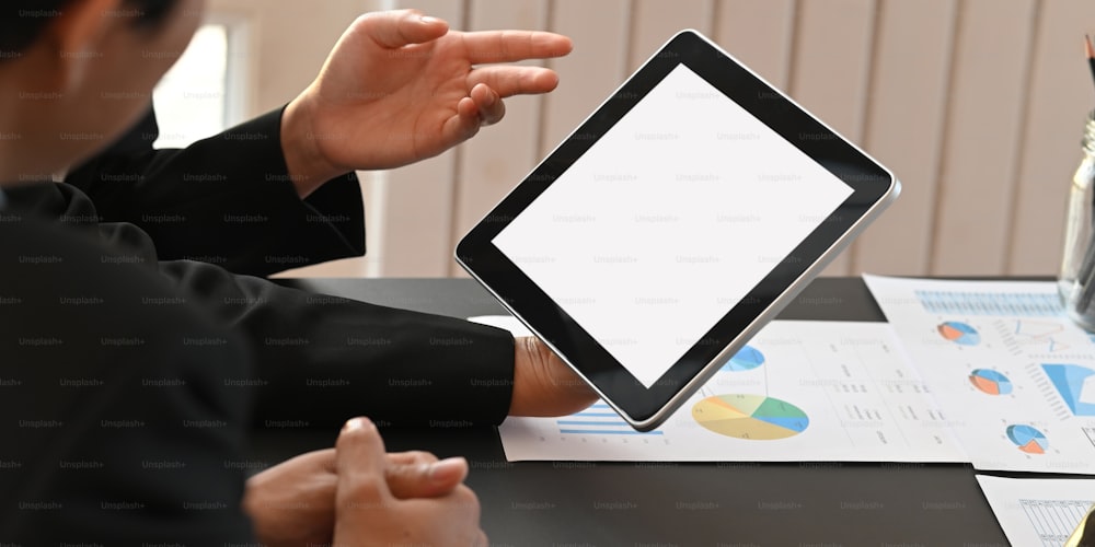 Cropped image of business people working together with computer tablet and graphic charts while sitting together at black meeting table over modern office as background.