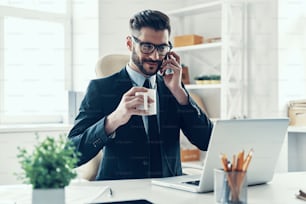 Happy young man in elegant business suit talking on the smart phone while sitting in the office