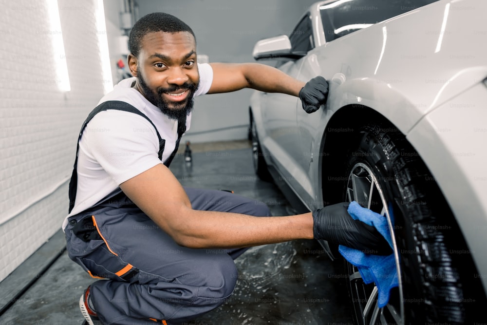 Manual car wash in car wash shop service. Handsome young dark skinned male employee worker cleaning the wheel rim of modern white car by blue microfiber cloth, looking at camera.