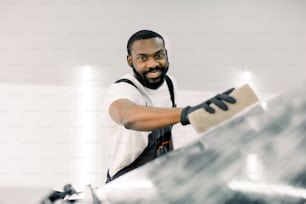 Smiling handsome African American man washing a soapy car windshield with a beige sponge, looking at camera. Car cleaning, wash service concept.