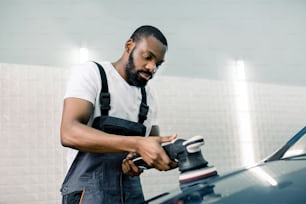 Servicio de detallado de coches. Guapo trabajador afroamericano barbudo con camiseta blanca y mono gris, sosteniendo una pulidora orbital en el taller de reparación mientras pule el coche. Enfoque selectivo