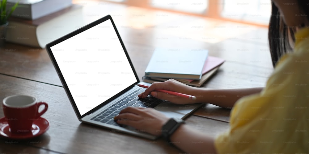 Cropped image of beautiful woman typing on computer laptop with white blank screen that putting on working desk and surrounded by notebook and coffee cup over comfortable living room as background.