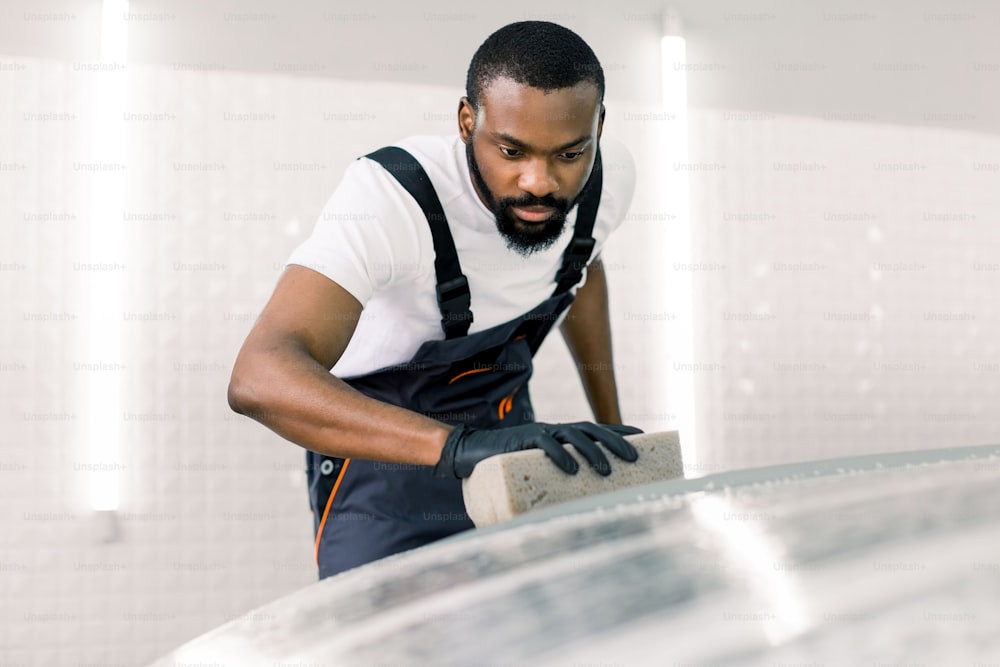 Joven trabajador masculino africano del servicio de automóviles con guantes de goma negros y mono gris, haciendo limpieza del capó del automóvil con la ayuda de champú y esponja amarilla en la empresa de lavado de autos.