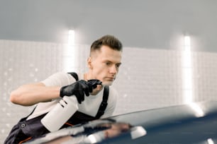 Close up of young car wash worker in special clothes and black rubber gloves, cleaning blue car hood with spray bottle,putting cold wax substance on surface. Car maintenance and detailing concept.