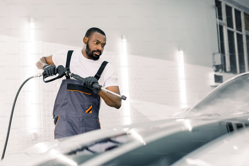 Limpieza del coche con pistola de agua a alta presión. Trabajador joven afroamericano guapo que lava automóviles de lujo modernos con agua a alta presión en un servicio profesional de lavado de autos.