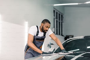 Young handsome African car wash worker man, wearing protective clothes and gloves, wiping modern car with microfiber cloth. Car detailing or valeting concept. Selective focus on face