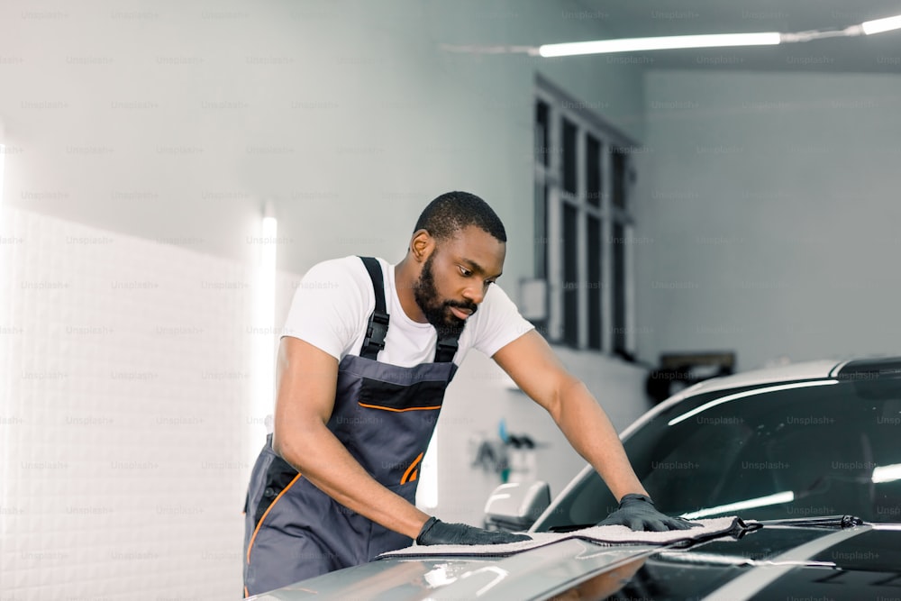 若いハンサムなアフリカの洗車労働者の男性は、防護服と手袋を着用し、マイクロファイバークロスで現代の車を拭いています。カーディテーリングまたはバレーのコンセプト。顔にセレクティブフォーカス