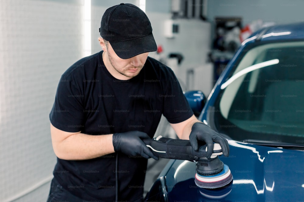Car detailing and polishing concept. Professional Caucasian male car service worker, wearing black t-shirt and cap, holding in hands orbital polisher, and polishing blue car in auto repair shop