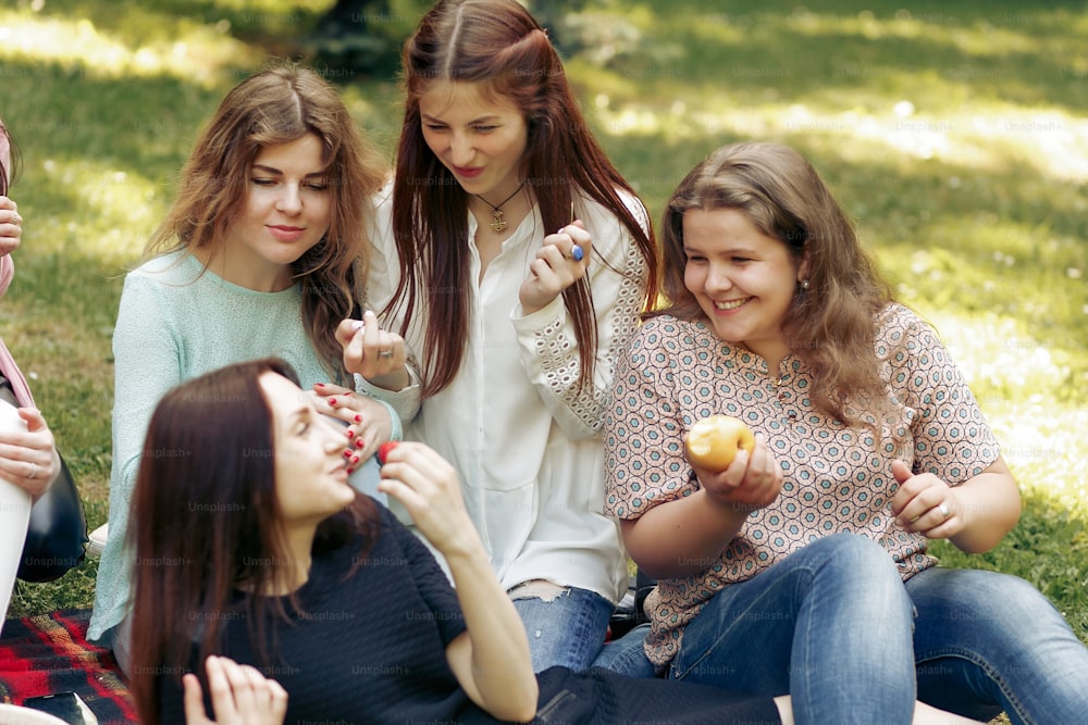 Gruppo felice alla moda di donne che mangiano frutta e si divertono sorridendo durante il picnic, momenti gioiosi nel parco estivo