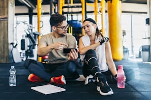 Male fitness gym instructor resting and sitting with young attractive woman on floor while making workout plan on his smart phone.