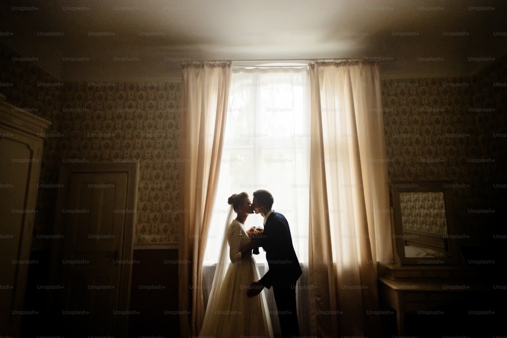 happy luxury bride and groom standing at window light in rich room, tender moment