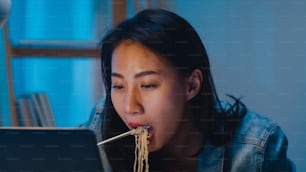 Freiberufliche kluge Geschäftsfrauen, die Instant-Nudeln essen, stressen sich, müde während der Arbeit am Laptop im Wohnzimmer zu Hause in der Nacht. Glückliches junges asiatisches Mädchen, das Überstunden am Schreibtisch sitzt, genießt die Entspannungszeit.