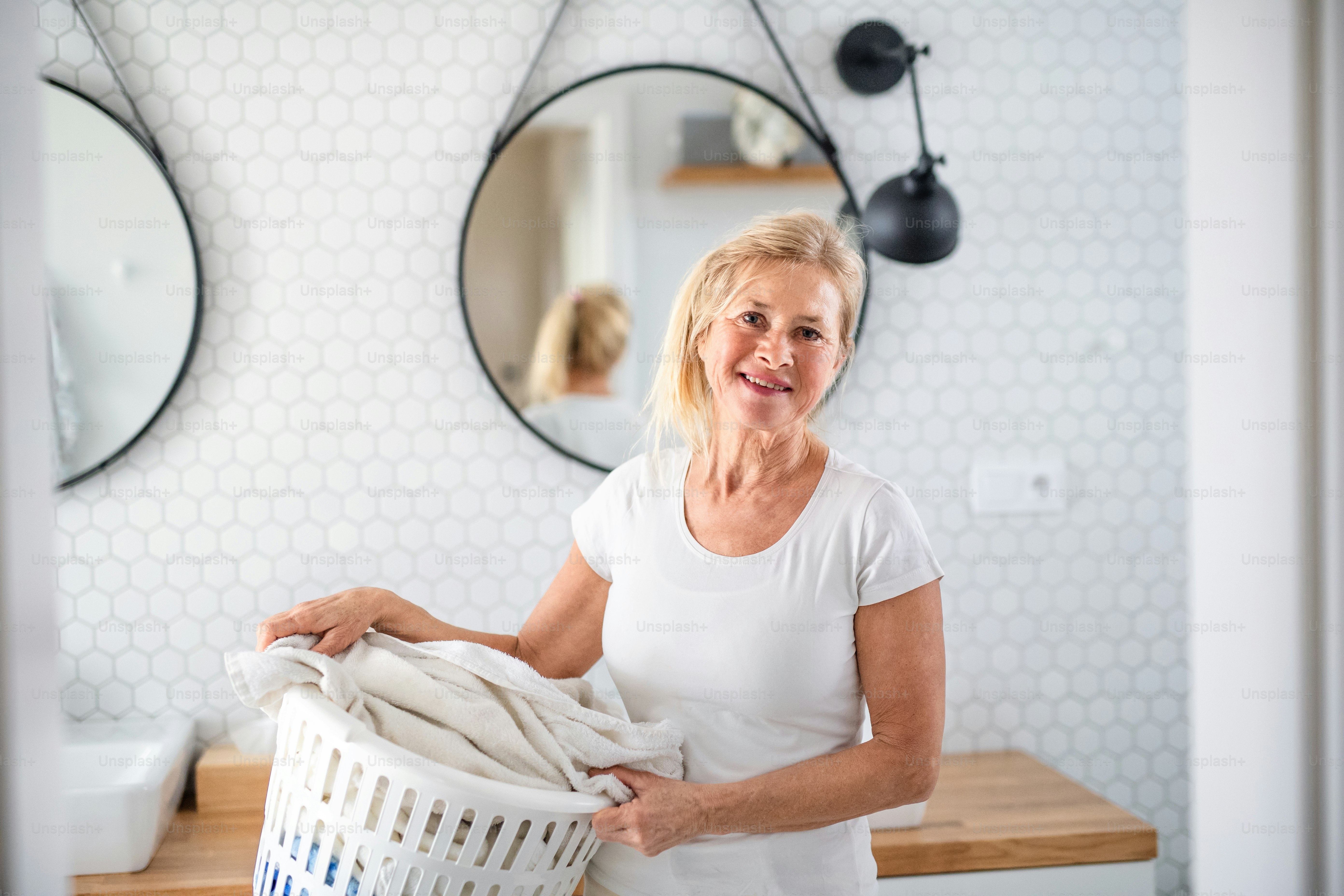 Retrato da mulher idosa com cesto de roupa no banheiro dentro de casa.