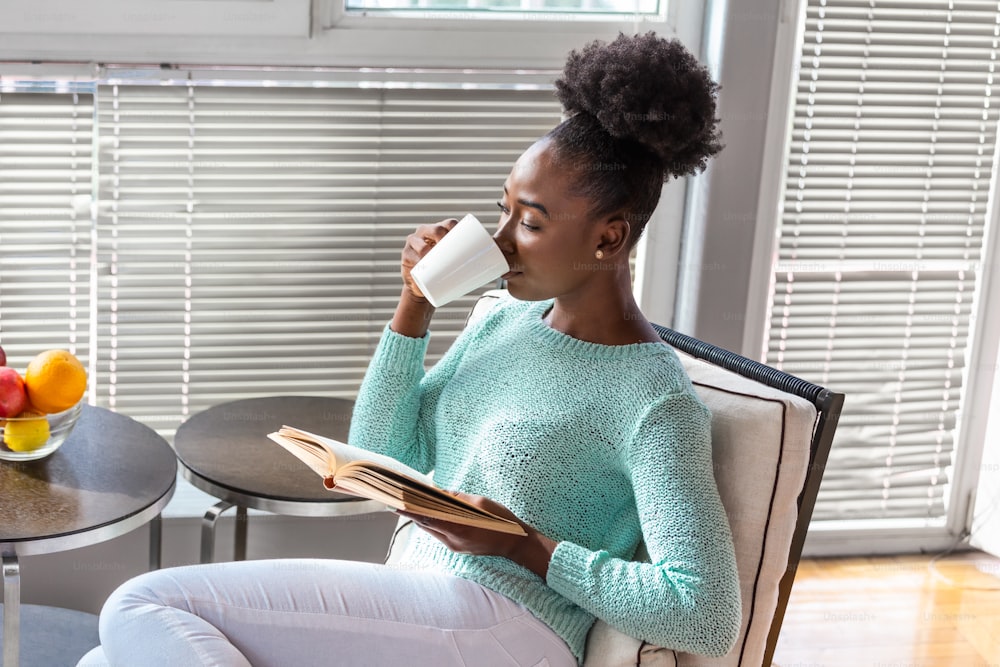 Young beautiful African American girl reading a book on the couch with the library bookshelves in the back. Beautiful woman on a white sofa reading a book