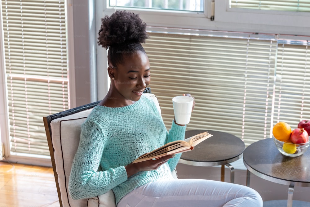 本と温かい飲み物のカップを持つ若い女性。本を読んでいるかなりアフリカ系アメリカ人の女の子は、奥に本棚のあるホームライブラリーに座ってコーヒーを楽しんでいます。