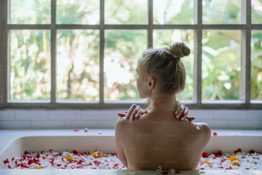 Organic skin care concept. Back view of young adult girl touching naked shoulders, lying in bathtub with herbs and flowers, resting in bathroom