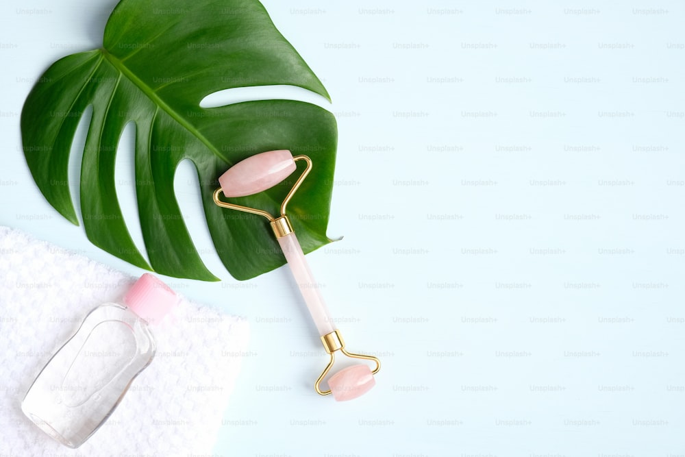 Rose quartz face roller and essential oil with monstera leaf on blue background. Flat lay, top view. Massage tool for facial skin care, SPA beauty treatment concept