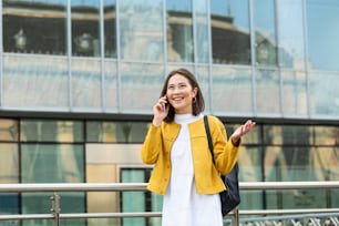 Cheerful young Asian woman walking along the street and calling on phone. Asian woman walking on the street, wearing cute trendy outfit and talking om her smart phone.