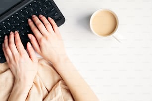 ノートパソコンのキーボードでテキストを入力する女性の手の上面図。フラットレイのモダンでミニマルな女性用ワークスペースに、ブランケット、コーヒーカップ、ノートを配