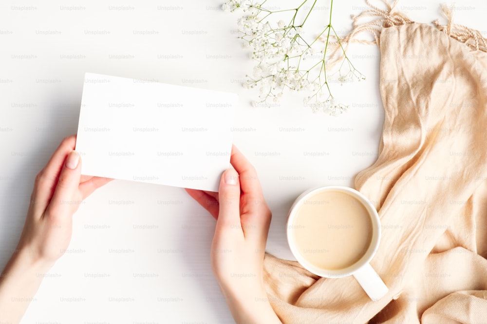 Manos femeninas sosteniendo una tarjeta de papel en blanco sobre un acogedor escritorio blanco de la casa con una taza de café, una manta, flores. Hygge, otoño otoño, concepto de confort. Plano plano, vista superior, espacio de copia