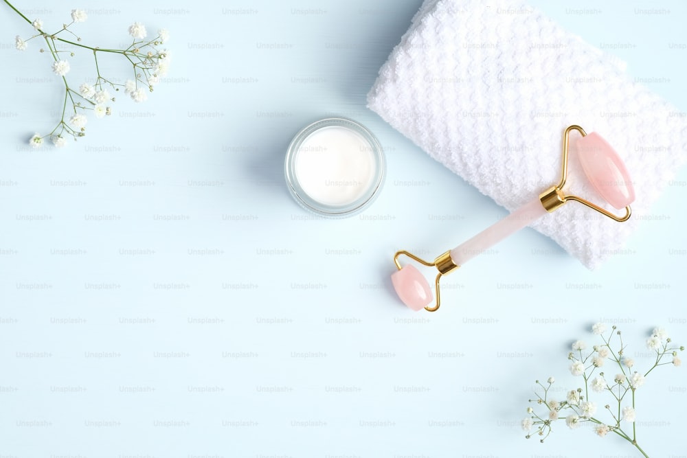 Rose quartz face roller for beauty facial massage therapy with moisturizer cream jar, towel and flowers on blue background. Flat lay, top view, copy space