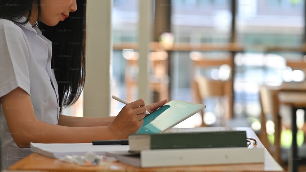 Image recadrée d’une femme médecin en blouse de laboratoire tenant un stylet et une tablette d’ordinateur dans les mains alors qu’elle est assise à un bureau en bois.