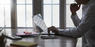Foto de homem inteligente na camisa branca manter a mão no queixo enquanto pensa e sentado na frente de um laptop de computador que coloca em mesa de trabalho de madeira sobre janelas confortáveis da sala de estar como fundo.