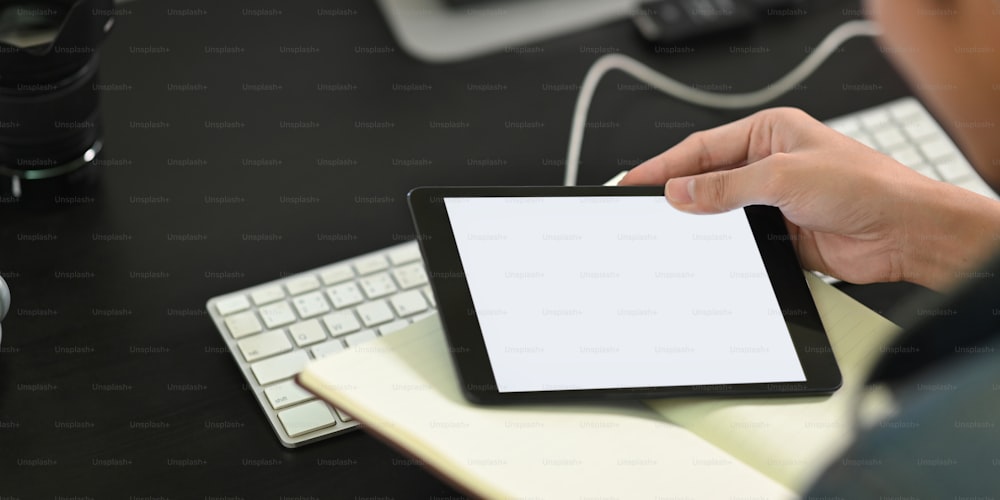Cropped image of man's hands holding a white blank screen computer tablet and notebook while sitting at the working desk over wireless keyboard and computer monitor as background.