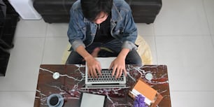 Imagem de visão superior do homem inteligente trabalhando como design gráfico enquanto se senta na mesa de textura de mármore e digita em seu laptop de computador sobre a confortável sala de estar como fundo.