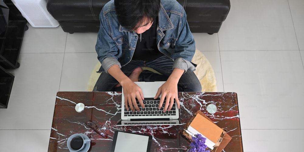 Top view image of smart man working as graphic design while sitting at the marble texture desk and typing on his computer laptop over comfortable living room as background.