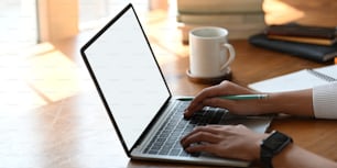 Abgeschnittenes Bild einer schönen Frau, die einen Computer-Laptop mit weißem leeren Bildschirm benutzt, während sie am weißen Schreibtisch über einem gemütlichen Wohnzimmer als Hintergrund sitzt.