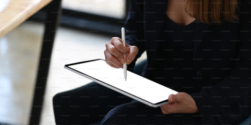 Cropped image of businesswoman holding a stylus pen while using a computer tablet and sitting at the wooden working desk over comfortable office as background.