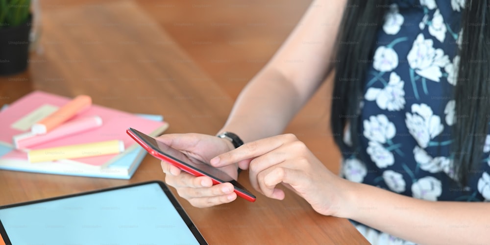 Cropped image of beautiful woman using a smartphone while sitting in front her computer tablet with white blank screen that putting on wooden working desk and surrounded by notebook and marker pens.