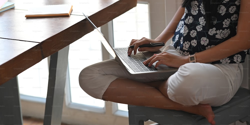 Image recadrée d’une belle femme tapant sur un ordinateur portable qui se met sur ses genoux tout en étant assise au bureau de travail en bois au-dessus d’un salon confortable en arrière-plan.